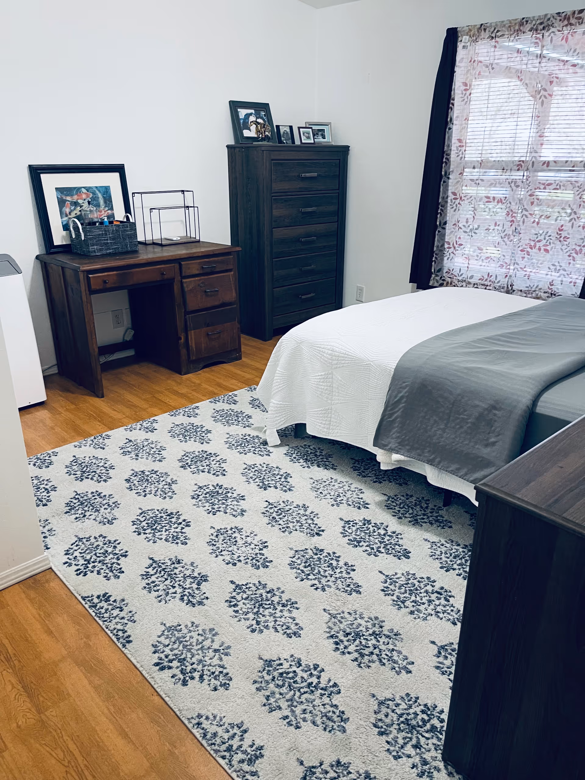 A tidy bedroom with a bed, dresser, tall chest, and a patterned rug.
