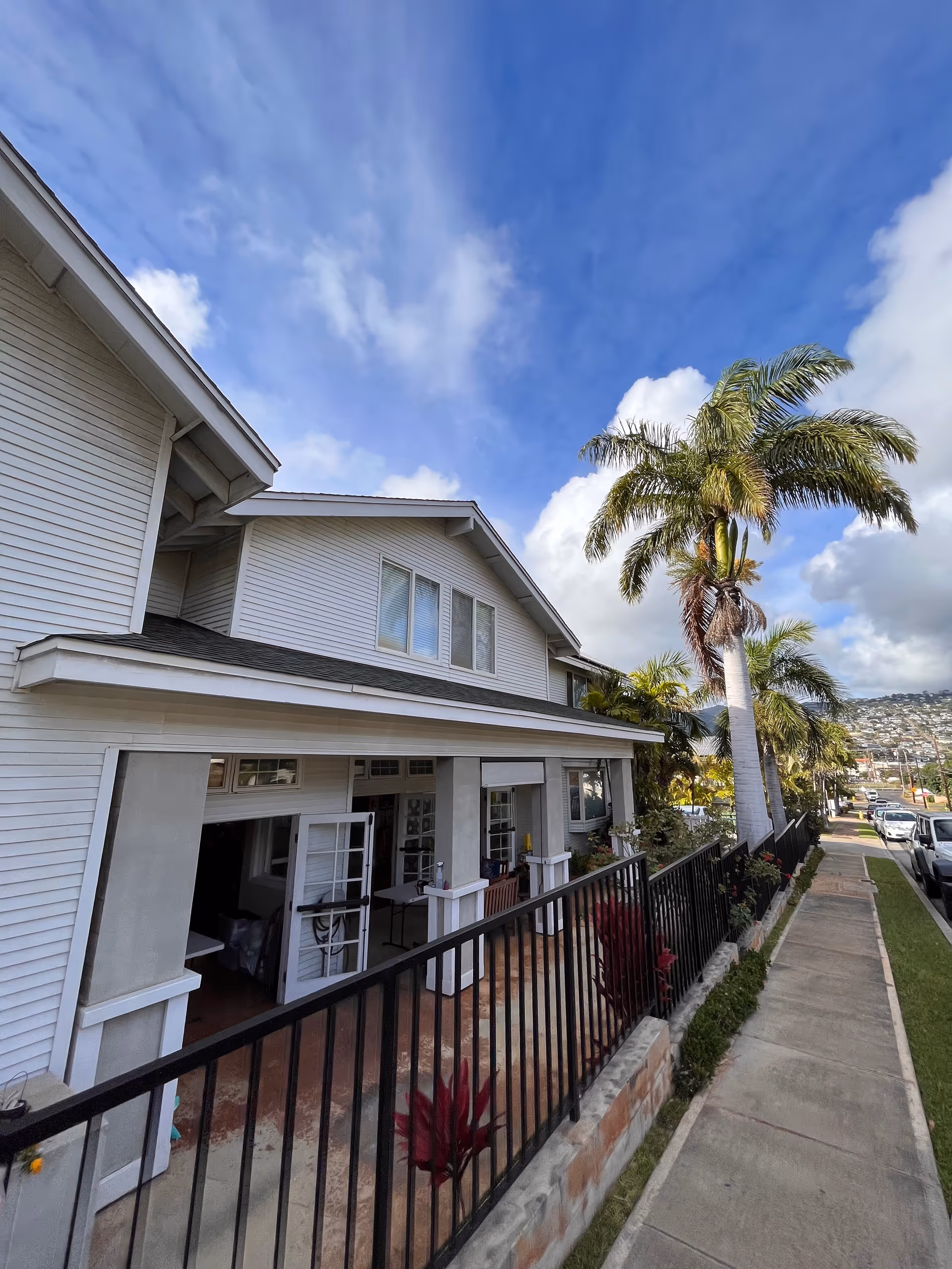 White residential-style senior care building exterior with a fenced porch, palm trees, and sidewalk.