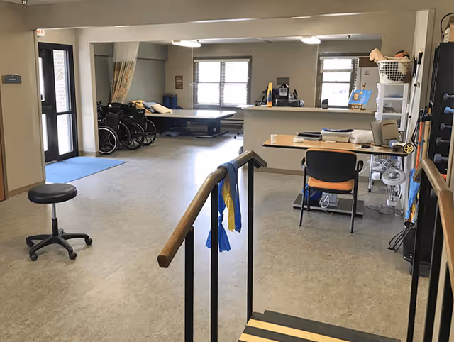 Interior activity area in a senior living facility with wheelchairs, a desk, and stair railing.