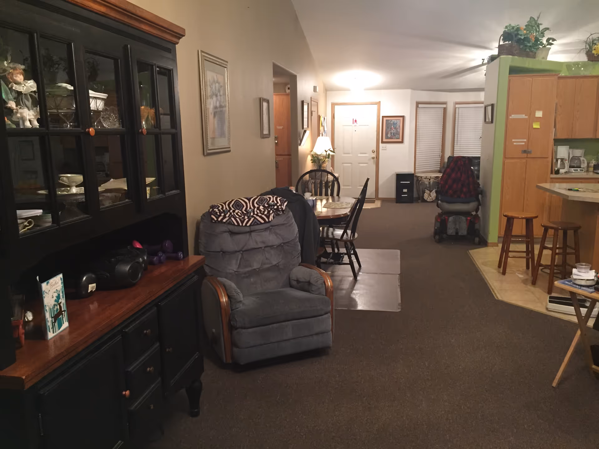 Cozy living room with dark cabinet, armchair, dining table, and a seated resident.