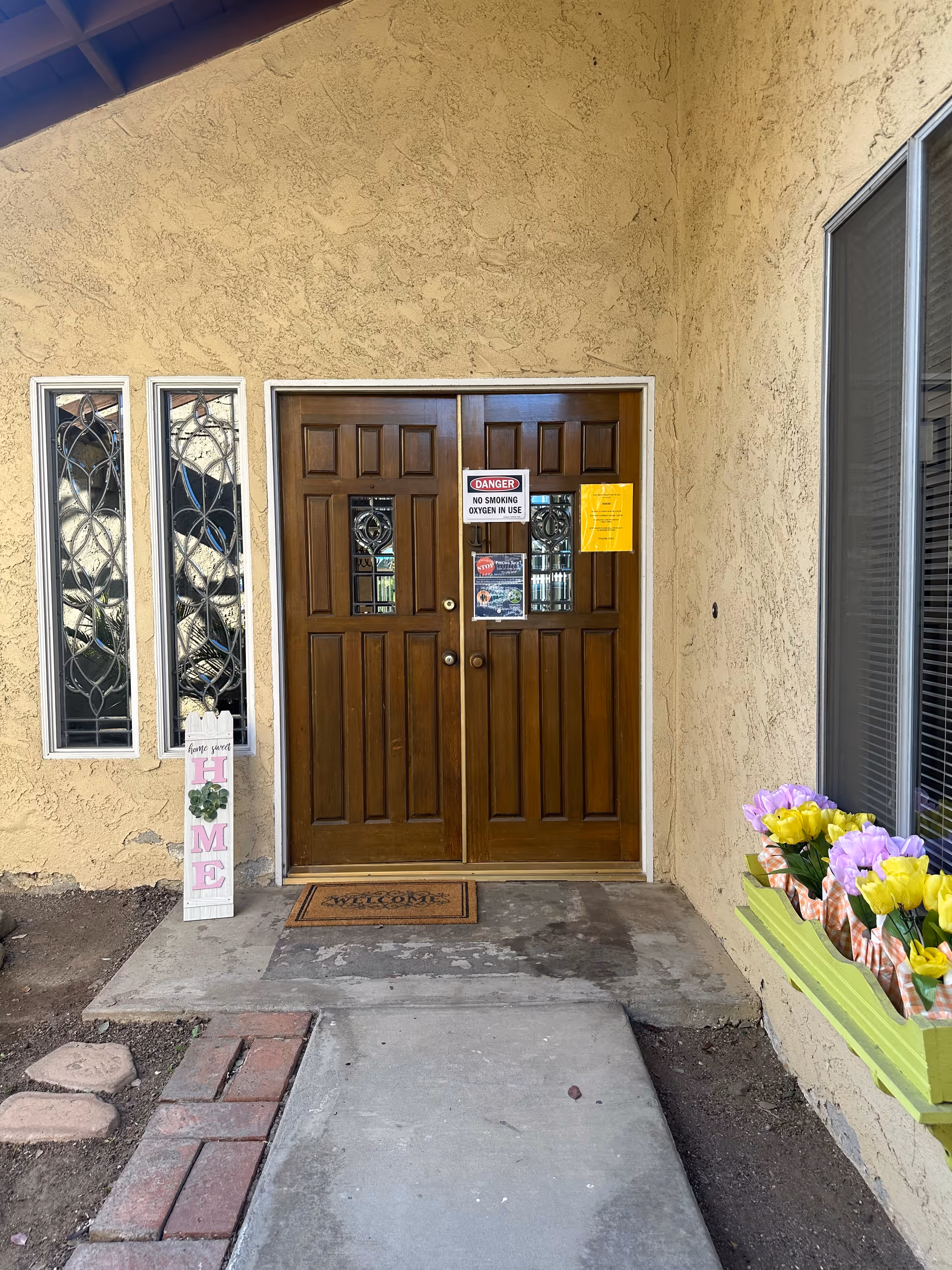 Double wooden front doors in a stucco entryway with a welcome mat, signs on the doors, decorative side windows, and a flower box with colorful tulips.
