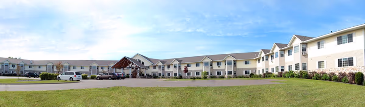 Wide exterior view of a two-story senior living facility building with beige siding, multiple windows, and a covered entrance. Several cars are parked in front of the building, and there is a large grassy area in the foreground under a blue sky with some clouds.