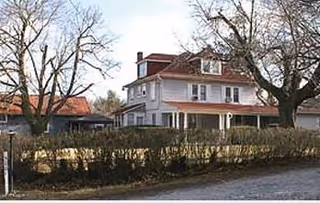 Two-story white building with red roof, front porch, trees, and hedges along the street.
