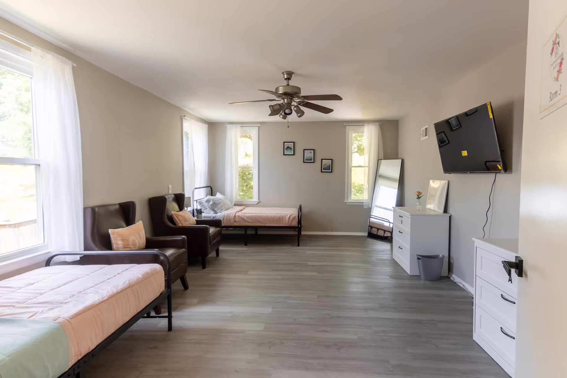 Bright senior living bedroom with two beds, chairs, dresser, mirror, and wall TV.