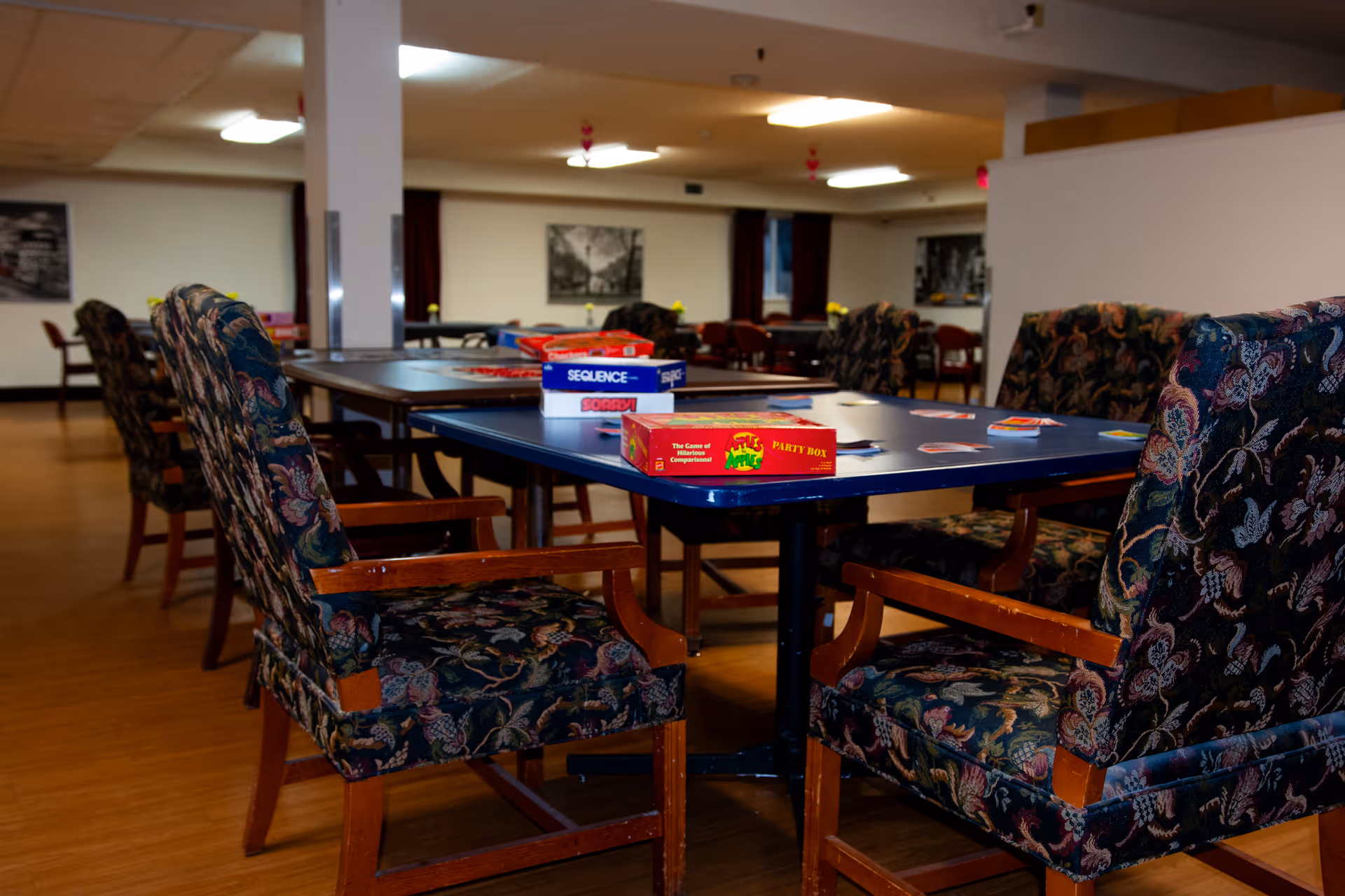A room with several tables and cushioned chairs arranged around them. On the tables are board games including Sequence, Sorry, and The Game of Things. The room has wooden flooring, white walls, and framed black and white pictures on the walls.