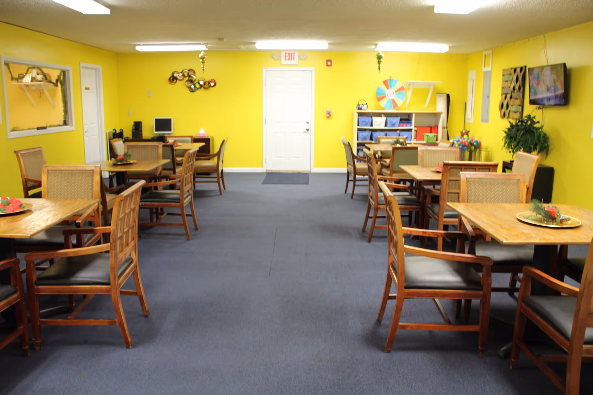 Bright yellow senior living dining room with several wooden tables, chairs, and decorative shelves.