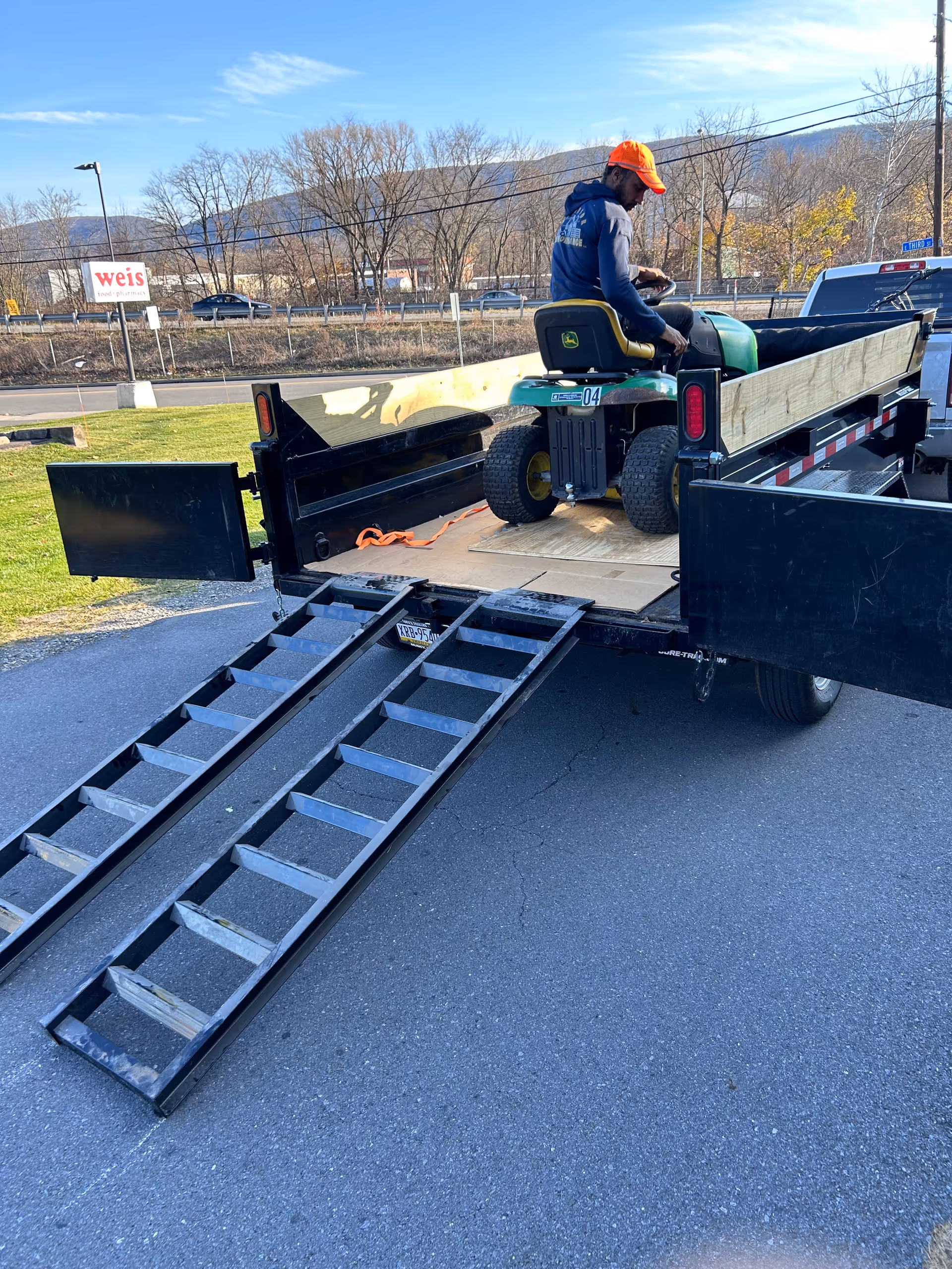 Man wearing orange cap sits on riding mower loaded on trailer outdoors near Williamsport