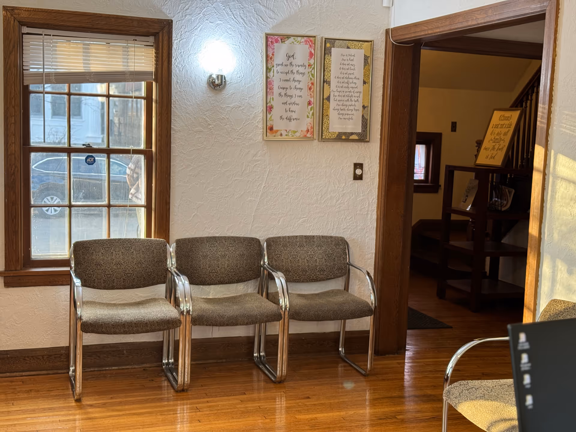 A small waiting area with three gray chairs along a textured white wall by a window.