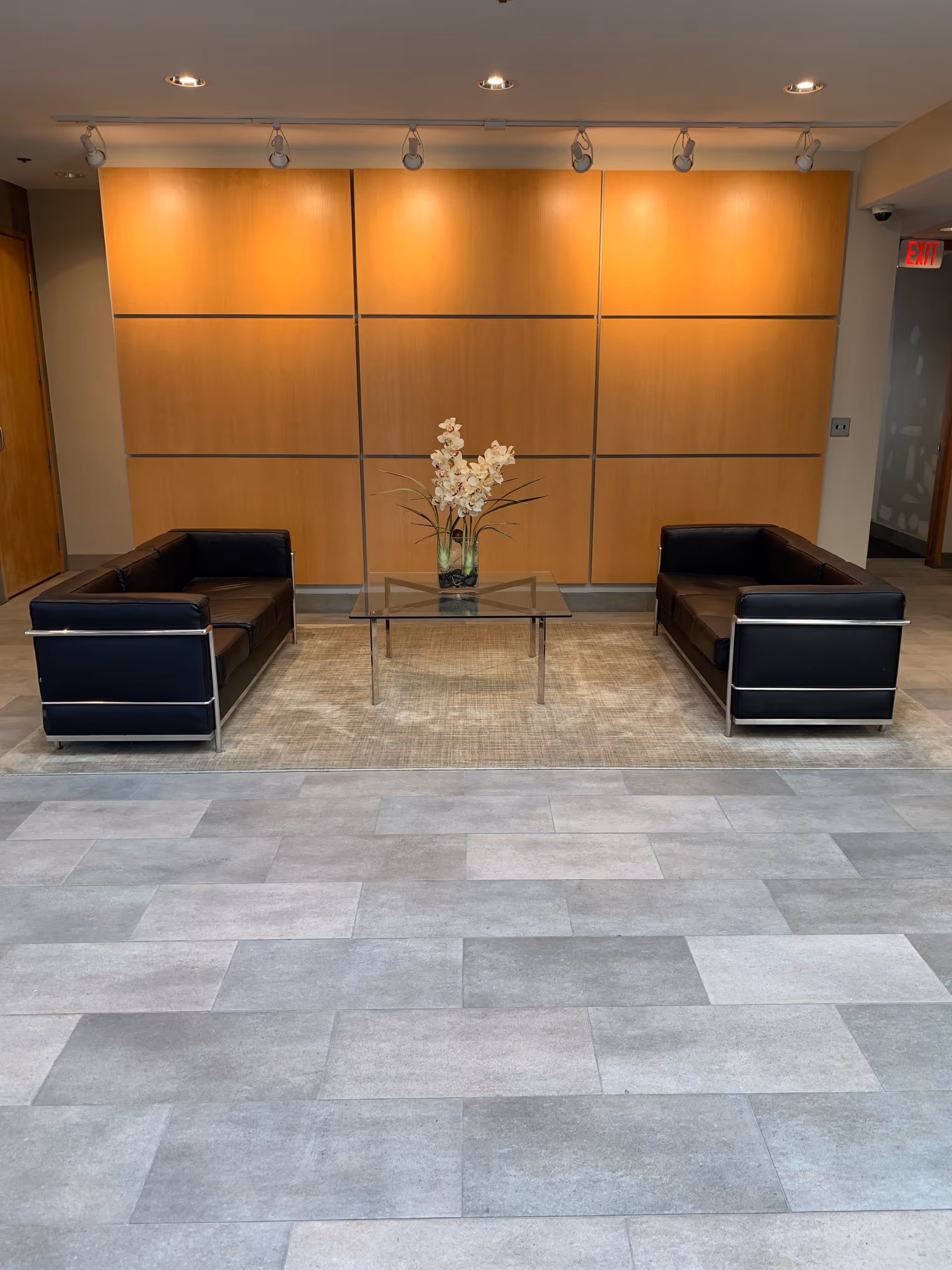 Modern lobby seating area with two black leather chairs, glass coffee table, and wood panel wall.