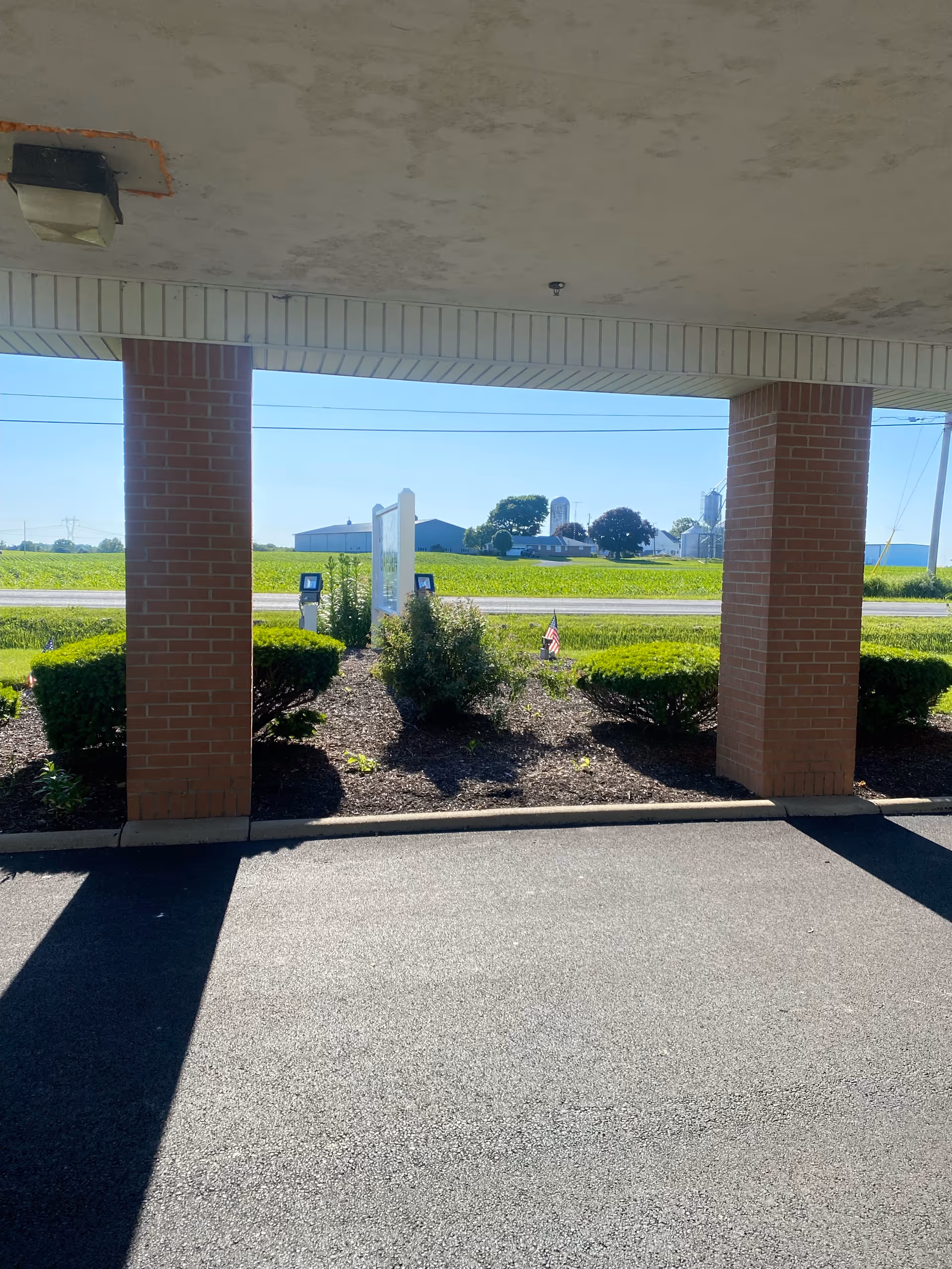 Covered driveway with brick columns, landscaped beds, and distant fields beyond a facility sign.