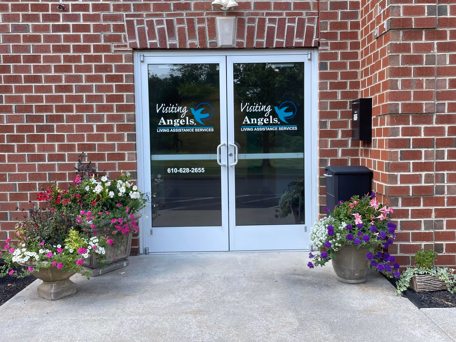 Glass doors with Visiting Angels signage flank a brick exterior, with colorful flower pots.