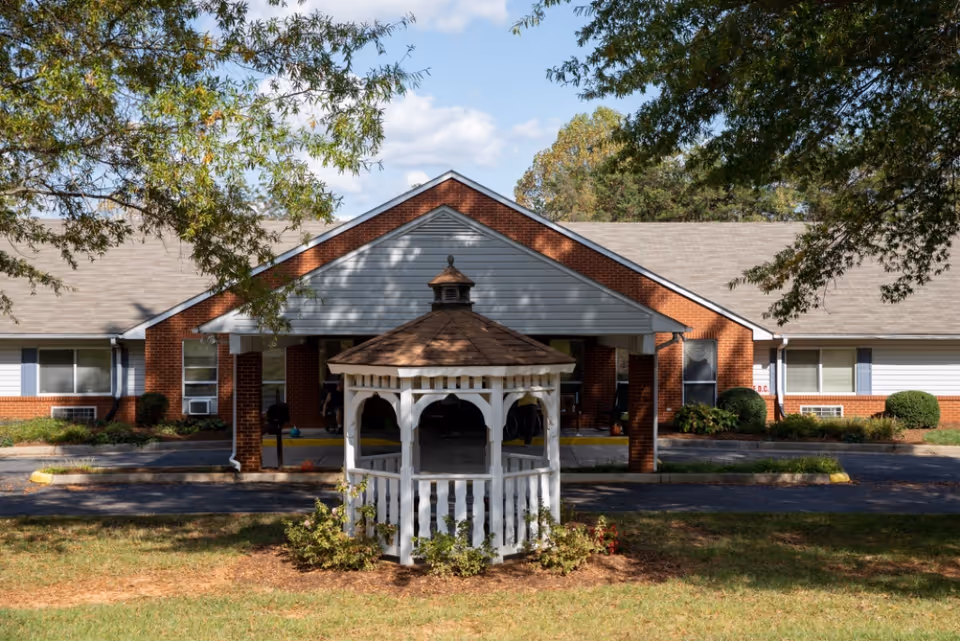 Front exterior view of a single-story brick and siding building with a covered entrance and a white gazebo in the foreground surrounded by grass and trees.