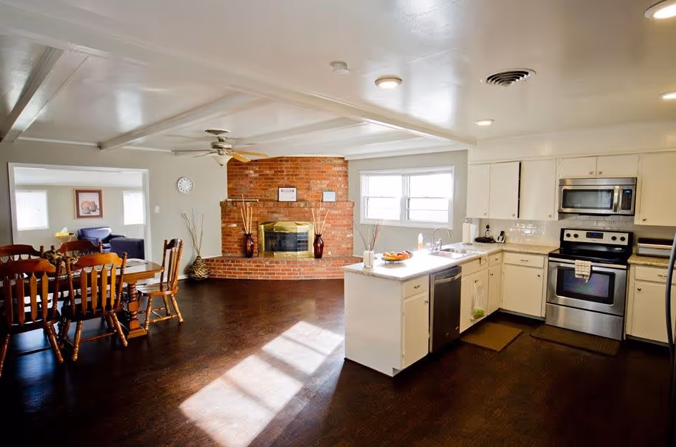 Bright open kitchen and dining area with white cabinets, stainless steel appliances, a kitchen island, wooden dining table with chairs, and a brick fireplace in the background. Adjacent living room area is partially visible.