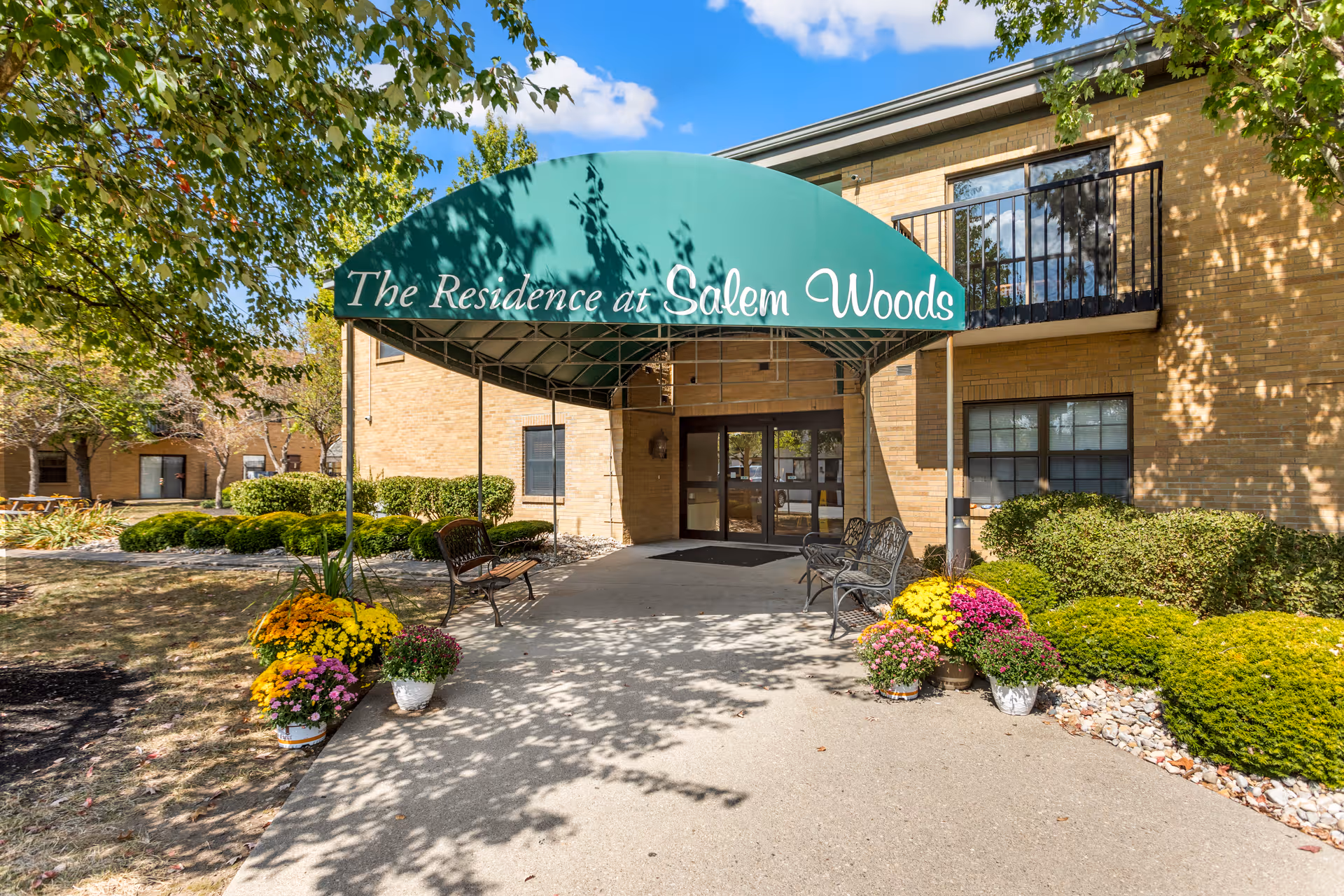 Entrance to The Residence at Salem Woods with a green canopy over the walkway, benches on either side, potted colorful flowers, and a brick building with windows and a balcony in the background.