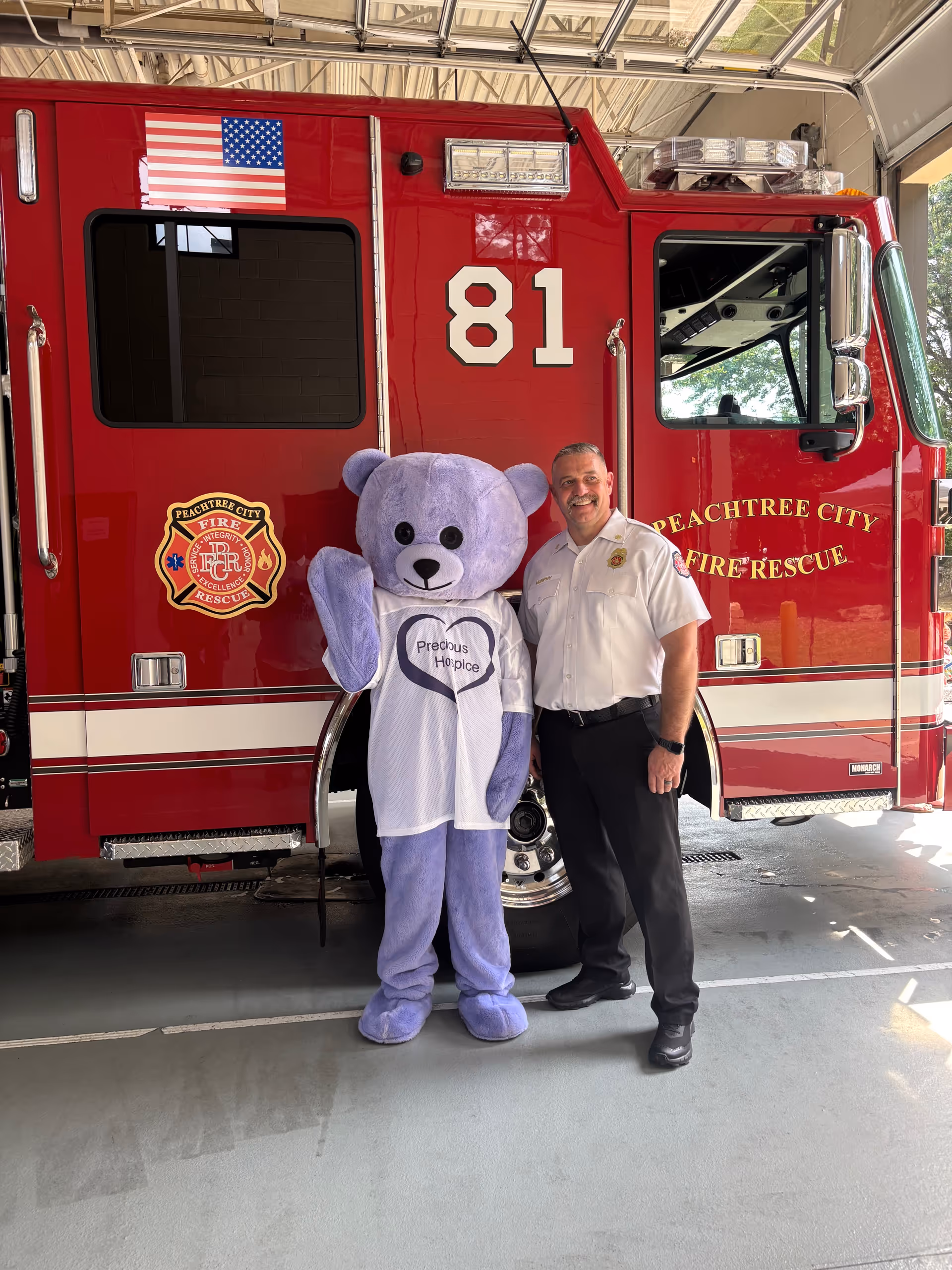 A purple teddy bear mascot and a firefighter stand beside a red fire truck indoors.