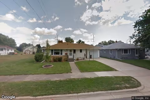 Single-story residential house with a front lawn, driveway, and a sidewalk in a suburban neighborhood under a partly cloudy sky.
