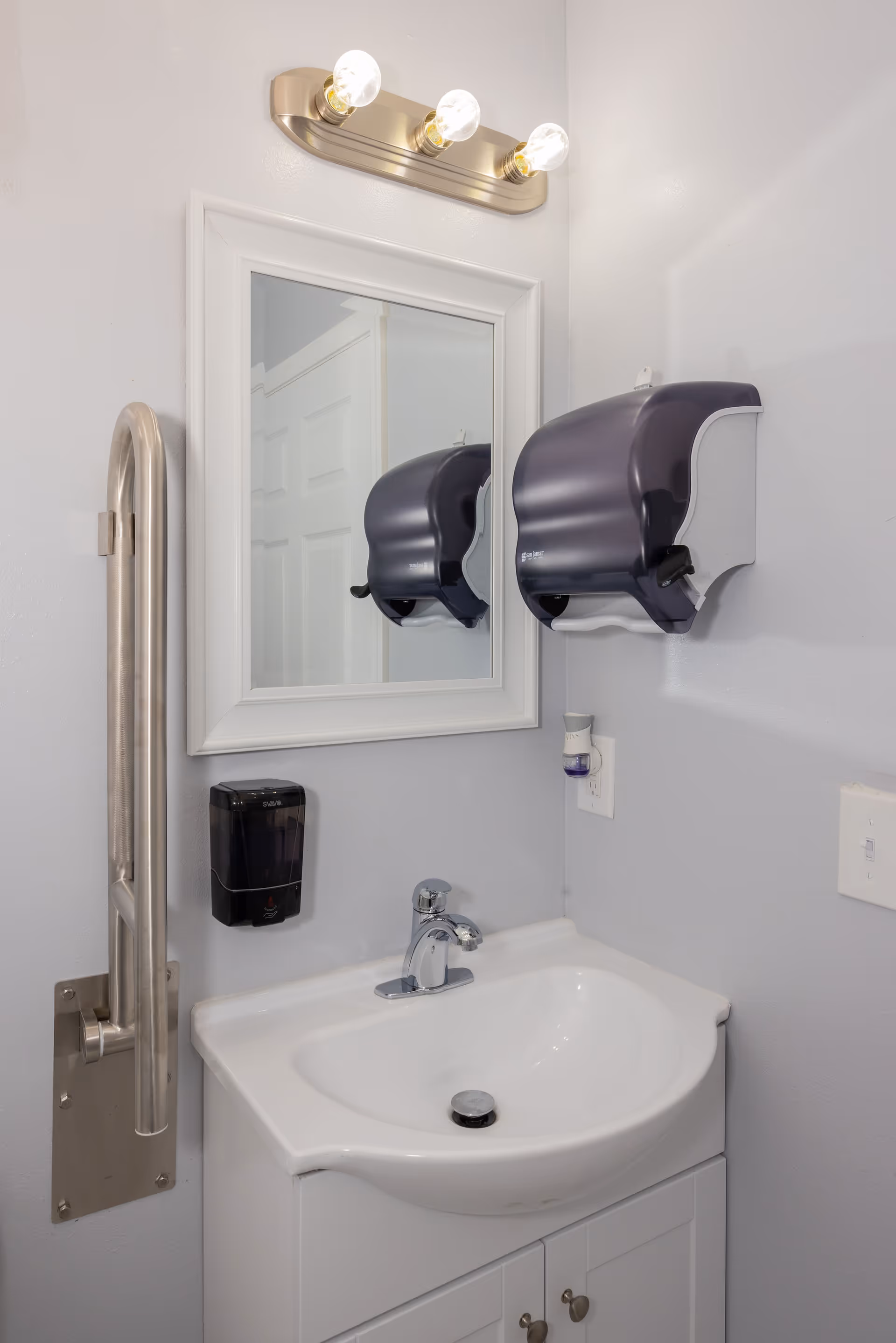 White bathroom with sink, mirror, soap dispenser, and overhead light.