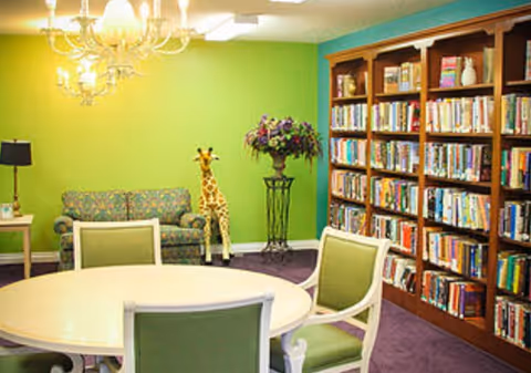 Bright common room with a round table and chairs, bookshelves, a couch and a decorative giraffe against a green wall.