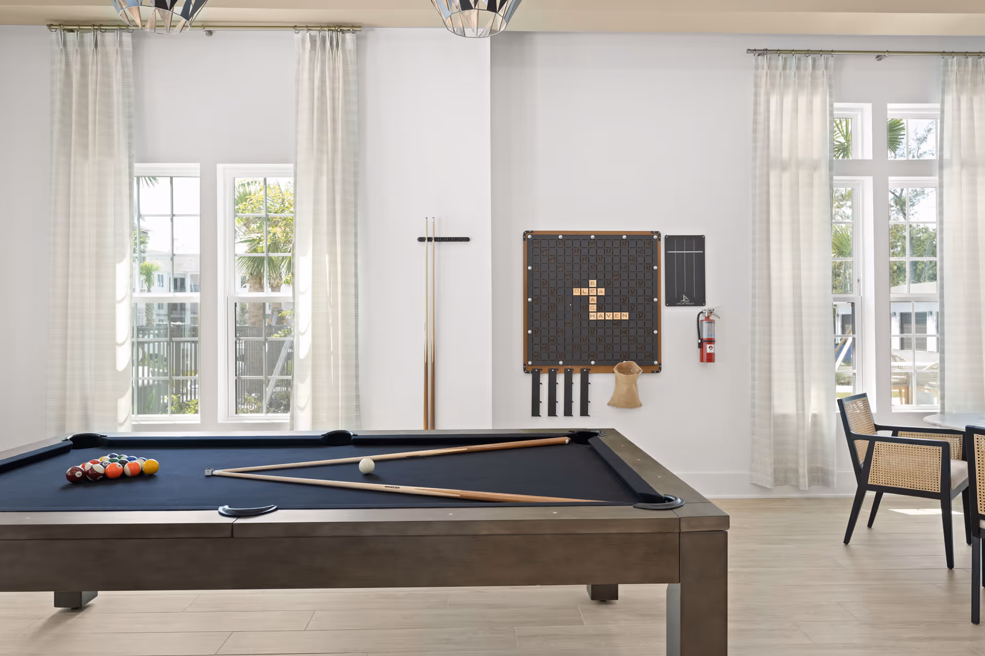 A bright and airy game room with a pool table in the center, pool cues resting on the table and on a wall rack, a large word game board on the wall spelling 'BEACH HAVEN', large windows with sheer white curtains letting in natural light, and a small seating area with chairs and a table near the windows.