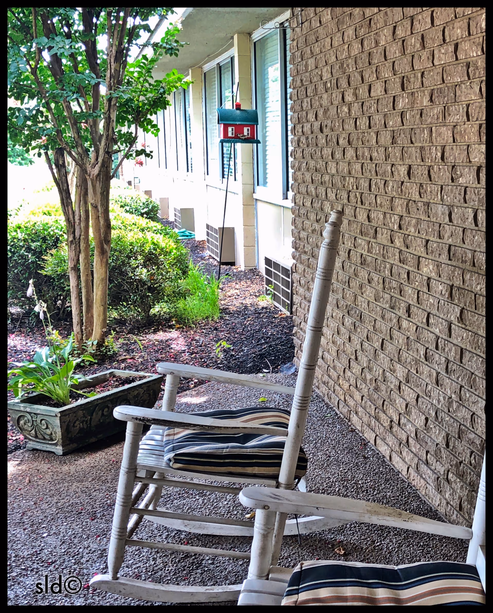 Outdoor porch with white rocking chairs, striped cushions, brick wall, and garden greenery.