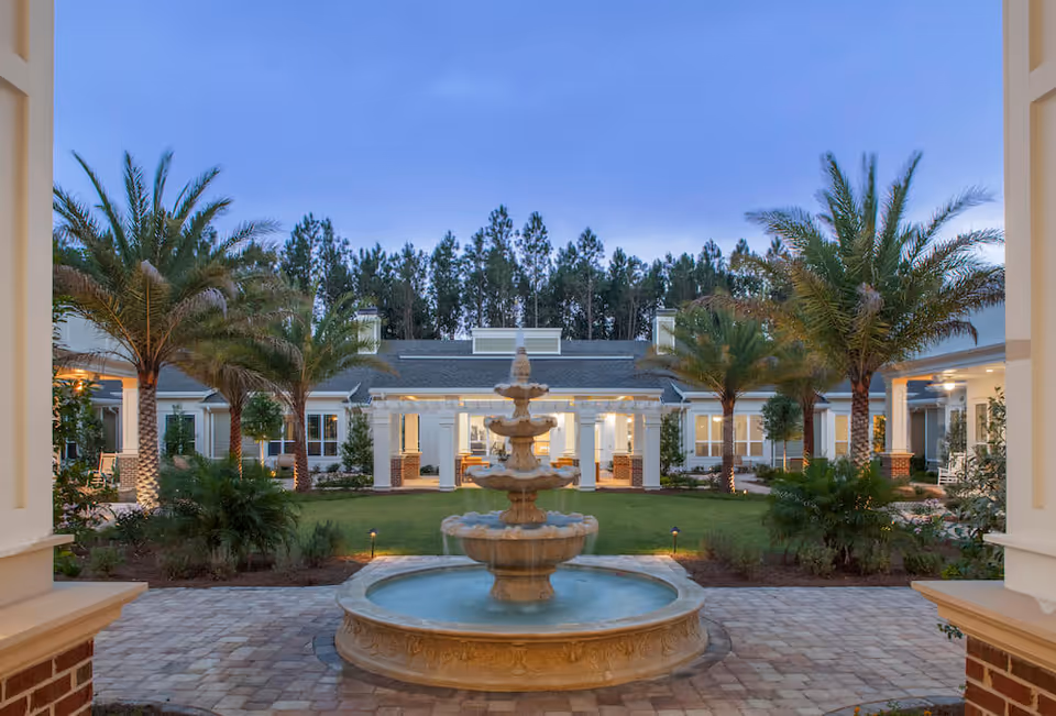 A central courtyard at dusk with a tiered fountain, palm trees, and single-story residence buildings surrounding a landscaped lawn.