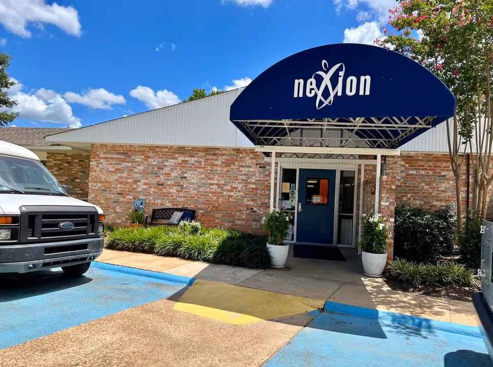 Front entrance of a brick building with a blue 'nexion' awning, double doors, potted plants, and a parked van.
