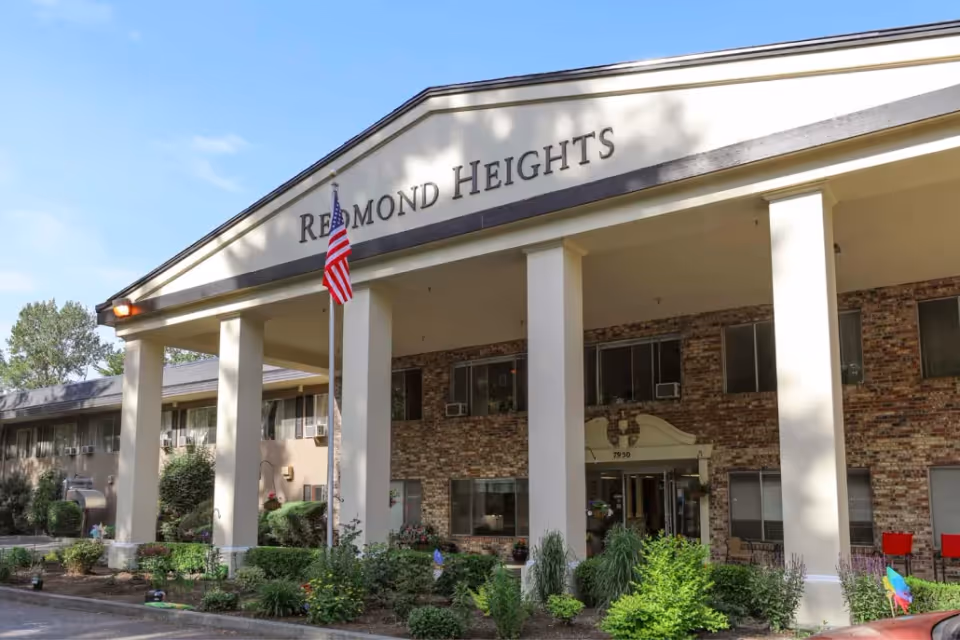 Front entrance of the Redmond Heights senior living building with tall columns, an American flag, and landscaping.