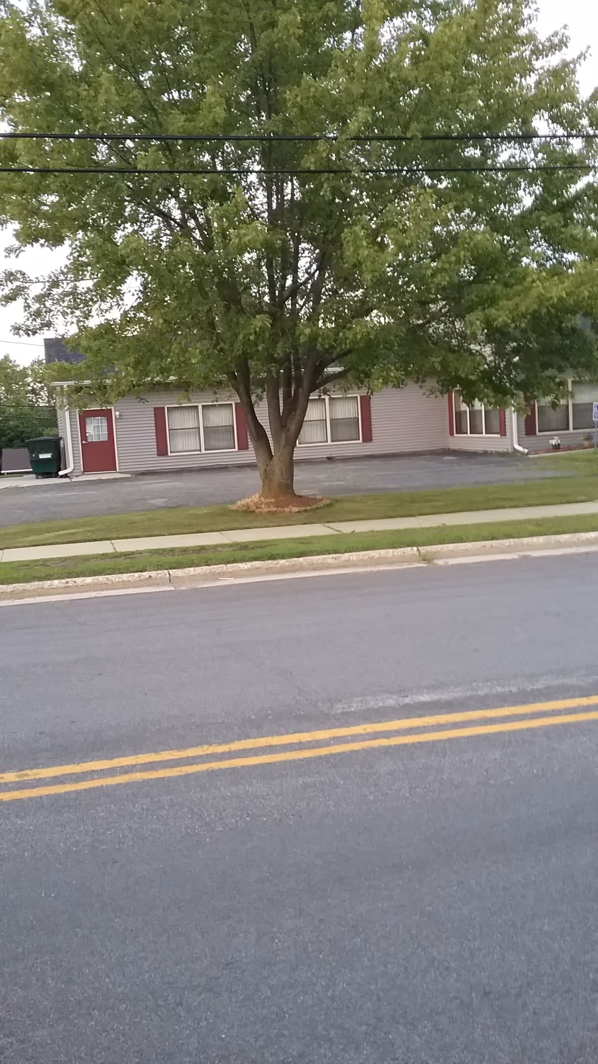 A single-story senior living facility exterior with a large tree, red shutters, and a street view.