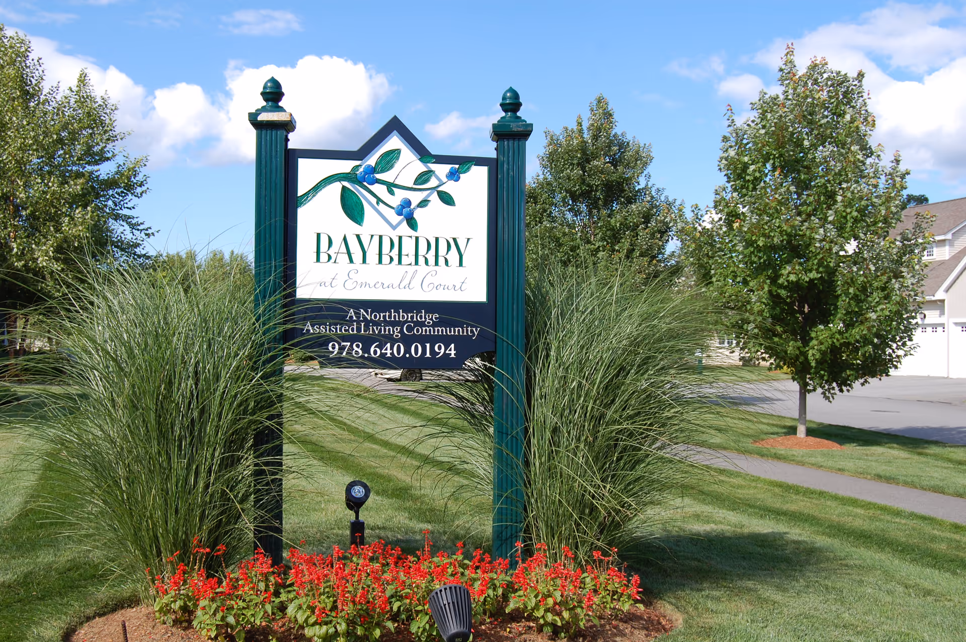 Outdoor view of a sign for Bayberry at Emerald Court, an assisted living community, surrounded by green grass, ornamental grasses, red flowers, and trees under a blue sky with some clouds.