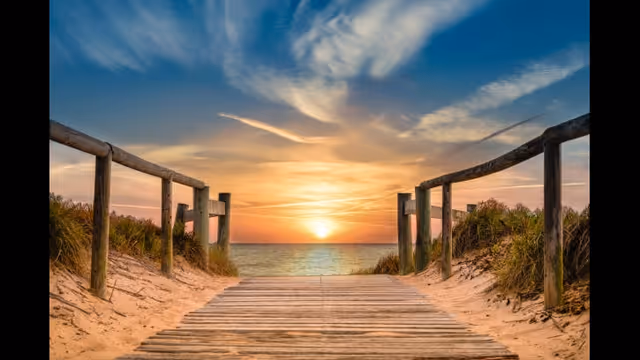Wooden boardwalk through sandy dunes toward a sunset over calm ocean.