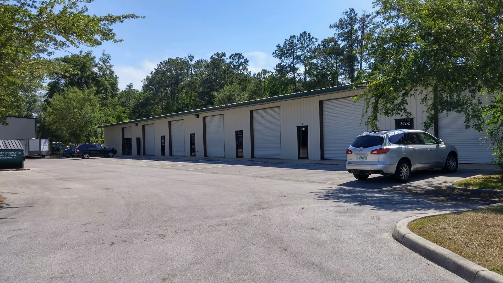 Exterior view of a long light-colored facility with large doors and a parking lot.