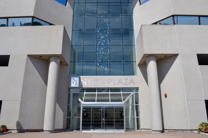 Modern building entrance with glass facade, concrete columns, and NYCB Plaza signage.