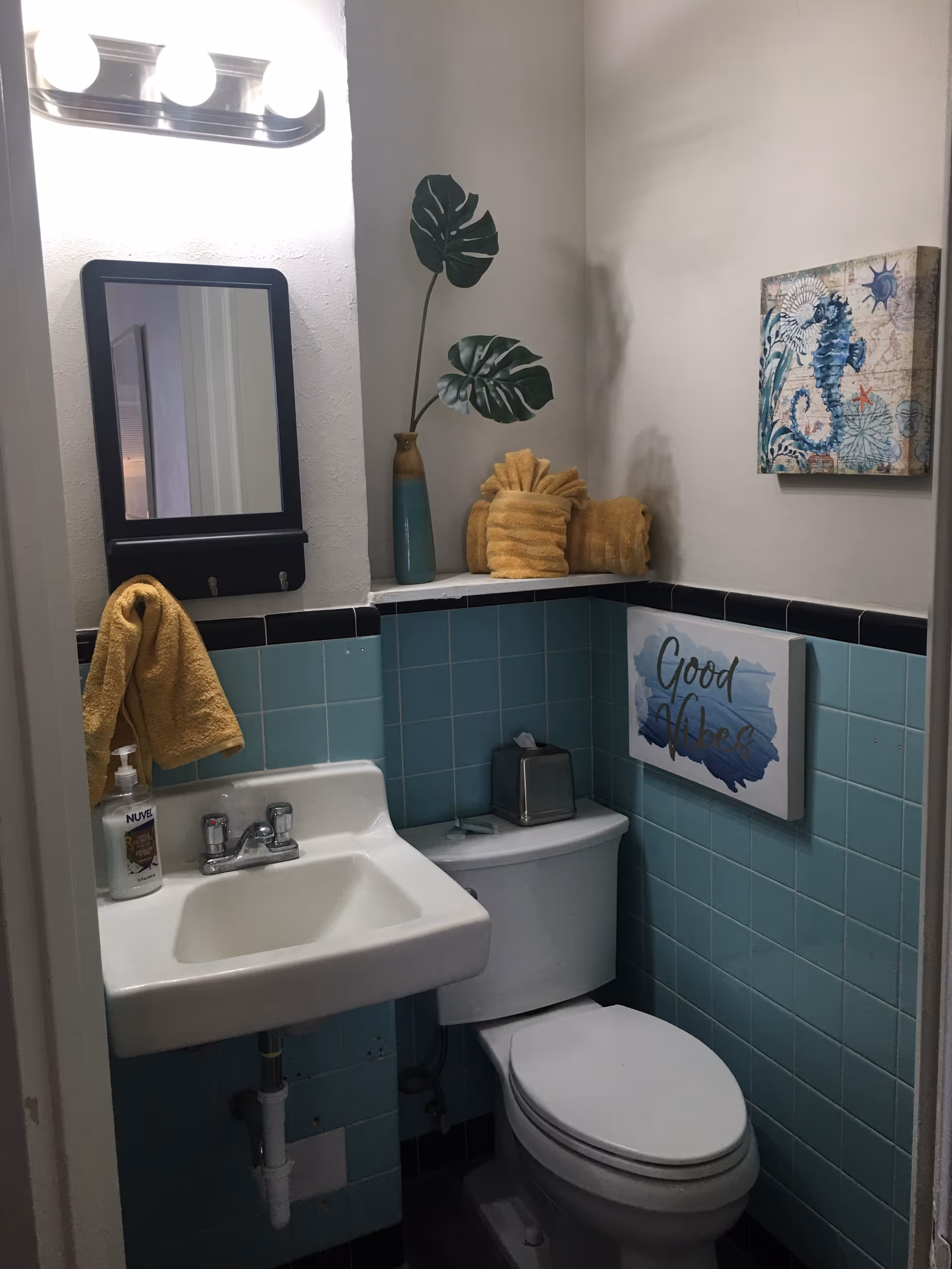 Small bathroom with turquoise tiled walls, white sink and toilet, mirror, towels, and coastal wall decor.