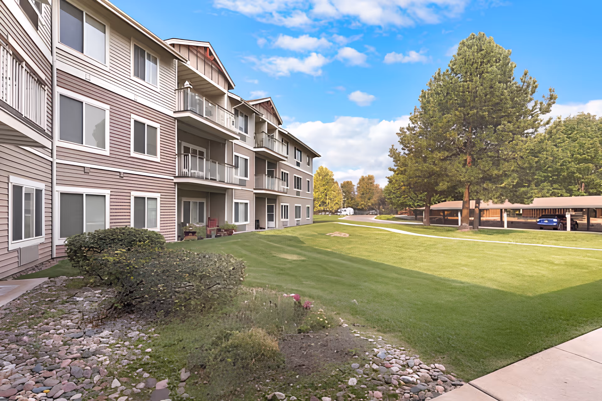 Exterior view of a multi-story residential building with balconies and windows, surrounded by a well-maintained lawn, bushes, trees, and a covered parking area under a blue sky with scattered clouds.