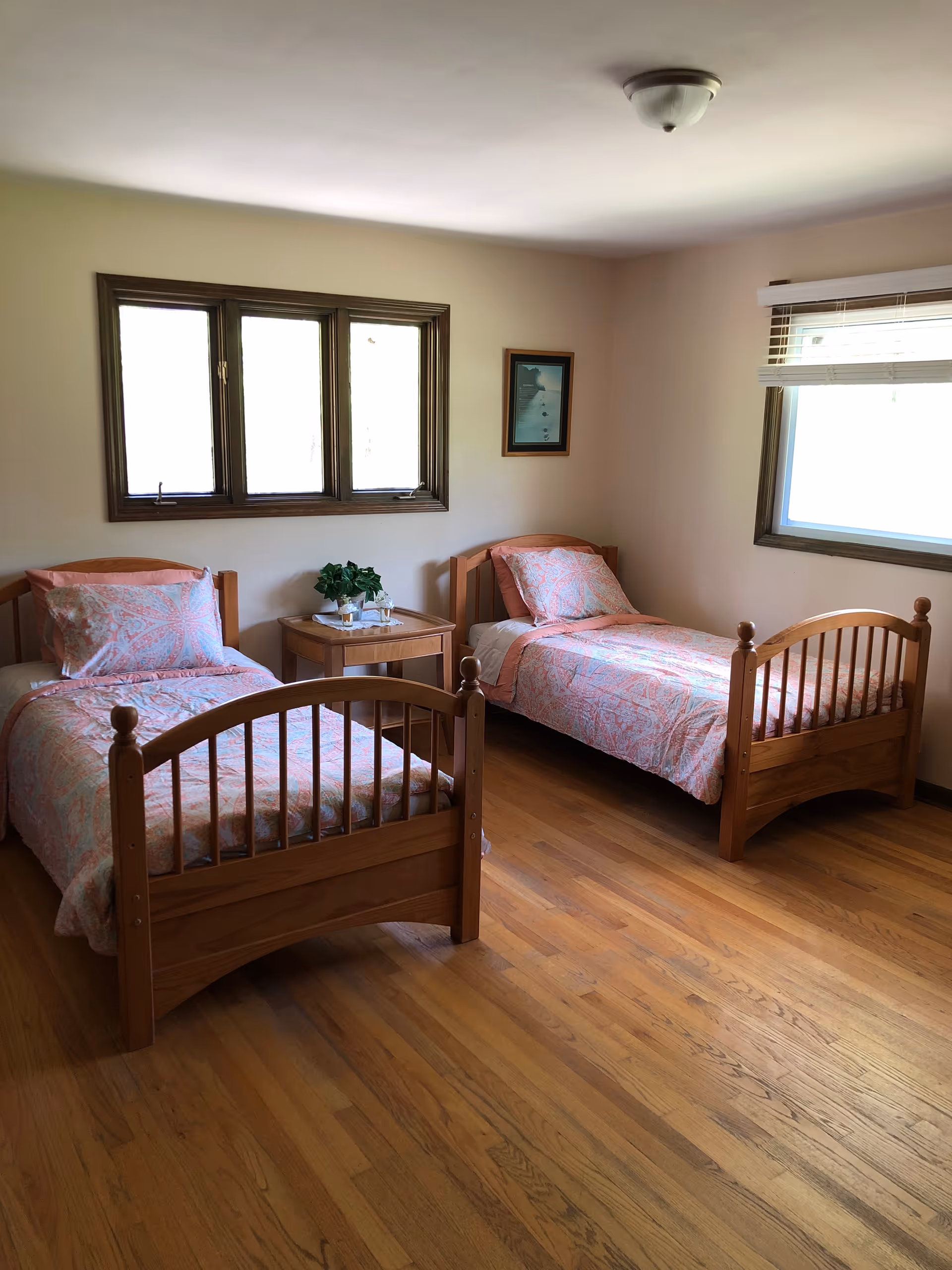 A simple, clean bedroom with two wooden twin beds, each made with matching pink and white patterned bedding. Between the beds is a wooden nightstand with a small green plant and two glasses of water. The room has hardwood floors, two windows with wooden frames, and a small framed picture on the wall.