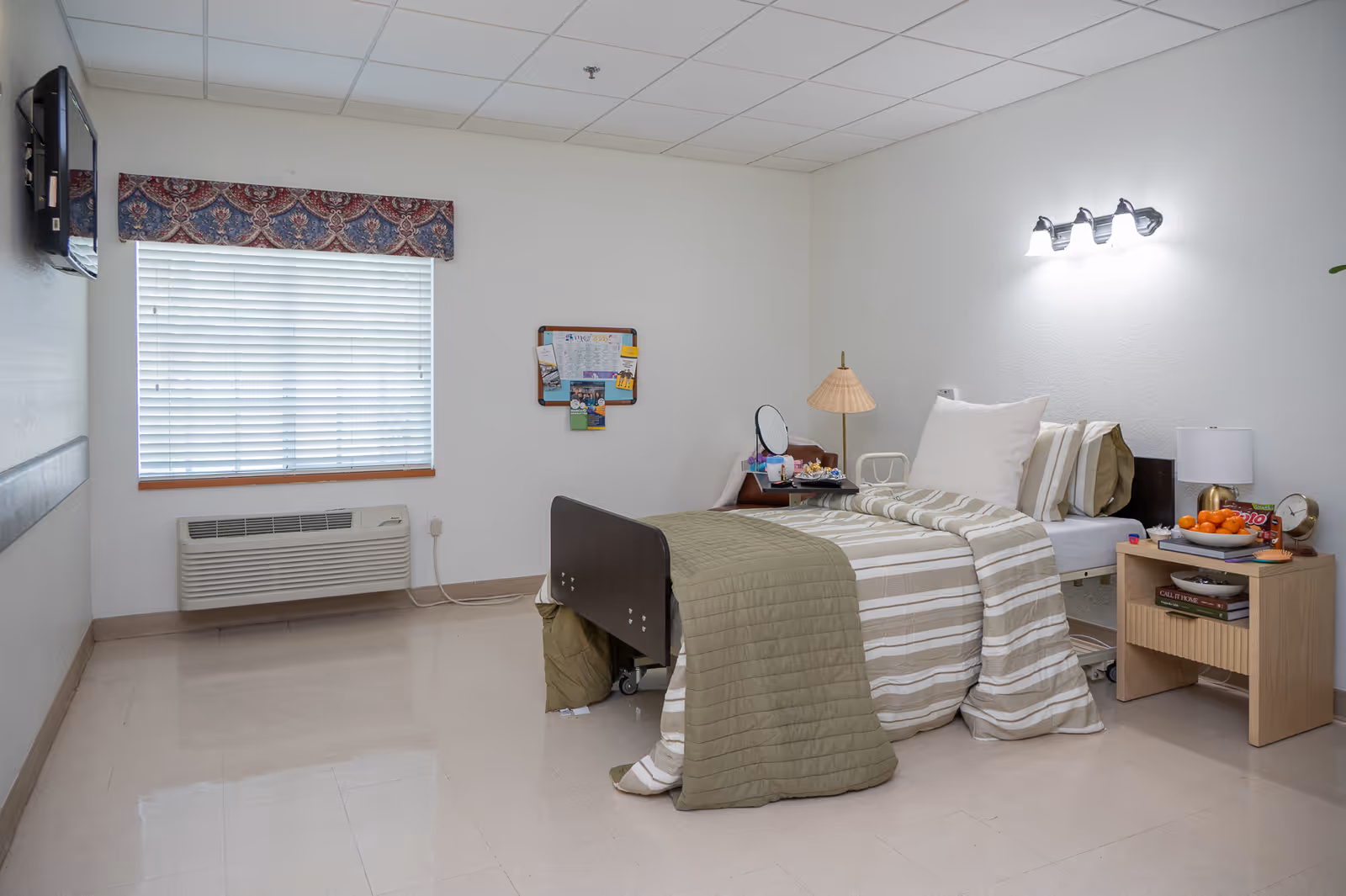A resident bedroom in a senior care facility featuring a bed, nightstand, and window.