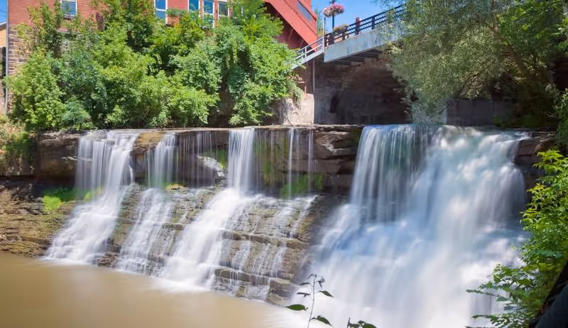Waterfall cascades beside a red-brick building with a pedestrian bridge overhead.