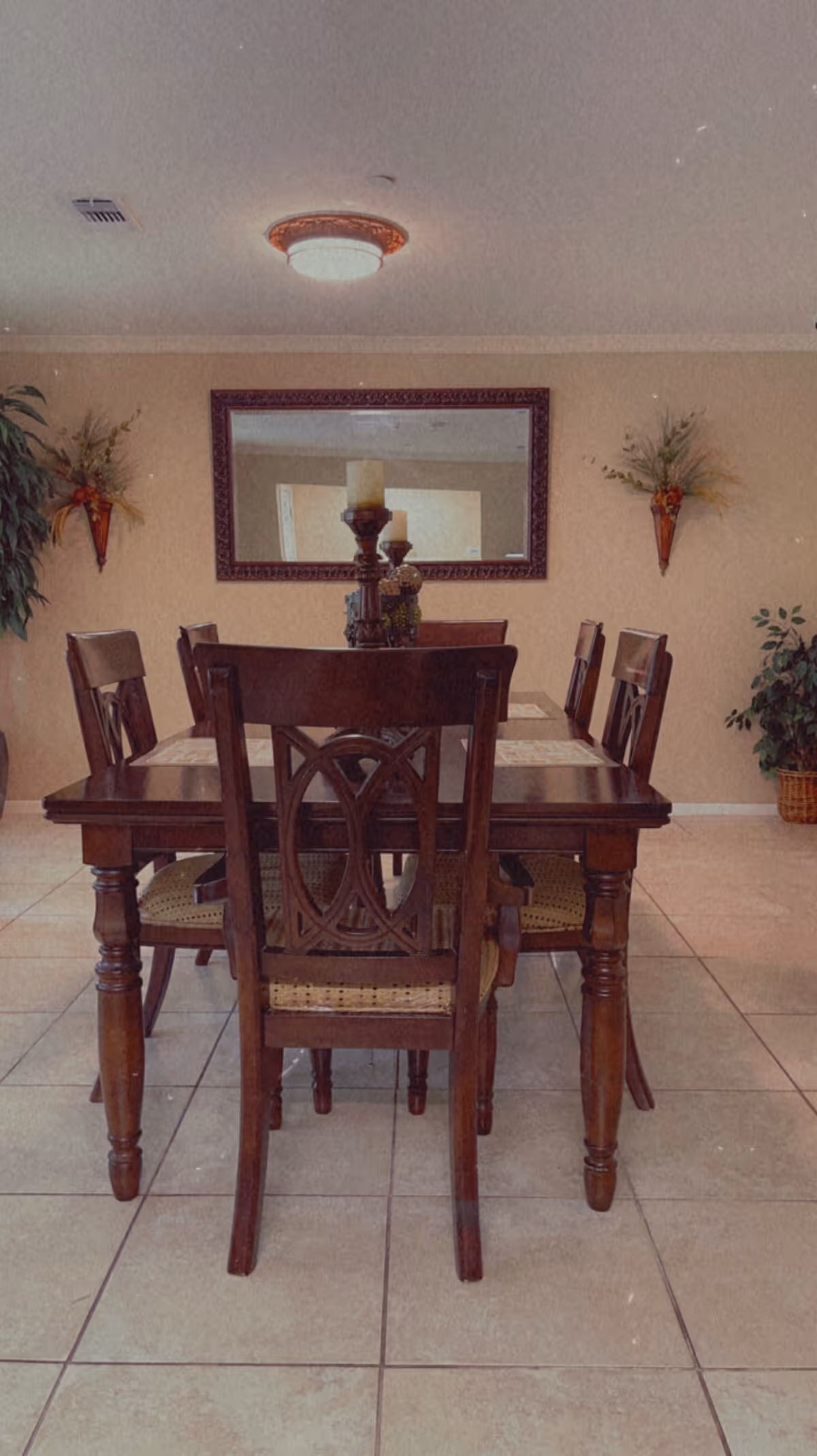 A dining room with a wooden dining table and six matching chairs. The table has a centerpiece with candles. There is a large rectangular mirror on the wall behind the table, flanked by two wall-mounted decorative planters with greenery. The floor is tiled, and there are additional potted plants in the corners of the room.