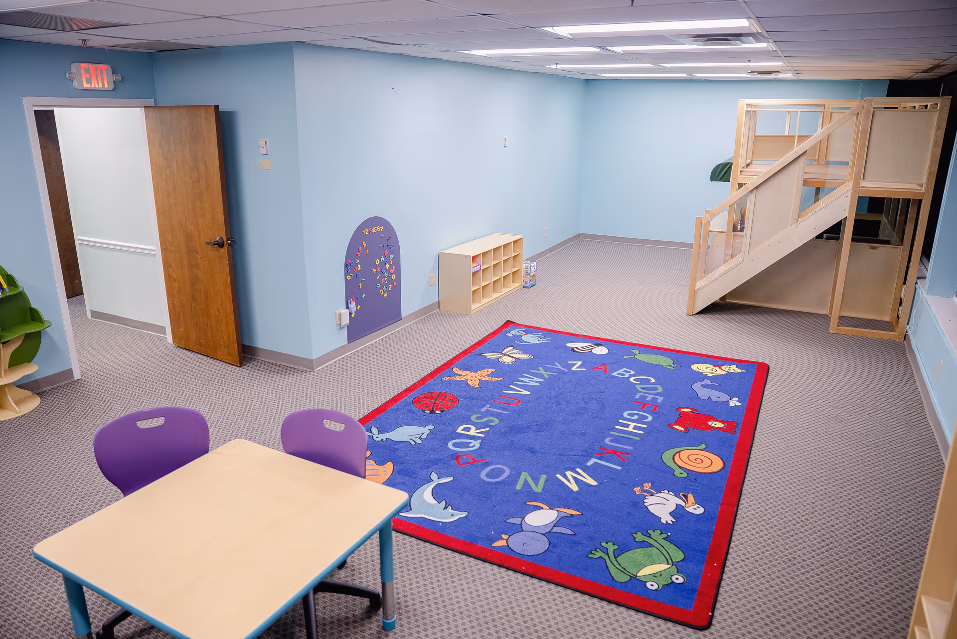 Blue therapy room with alphabet rug, small table, purple chairs, and a lofted play structure.