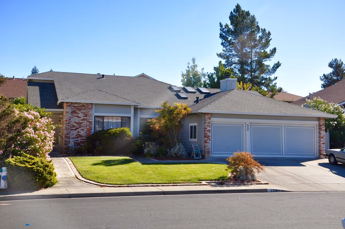 Single-story suburban house with a three-car garage, manicured front lawn, and trees under a clear blue sky.