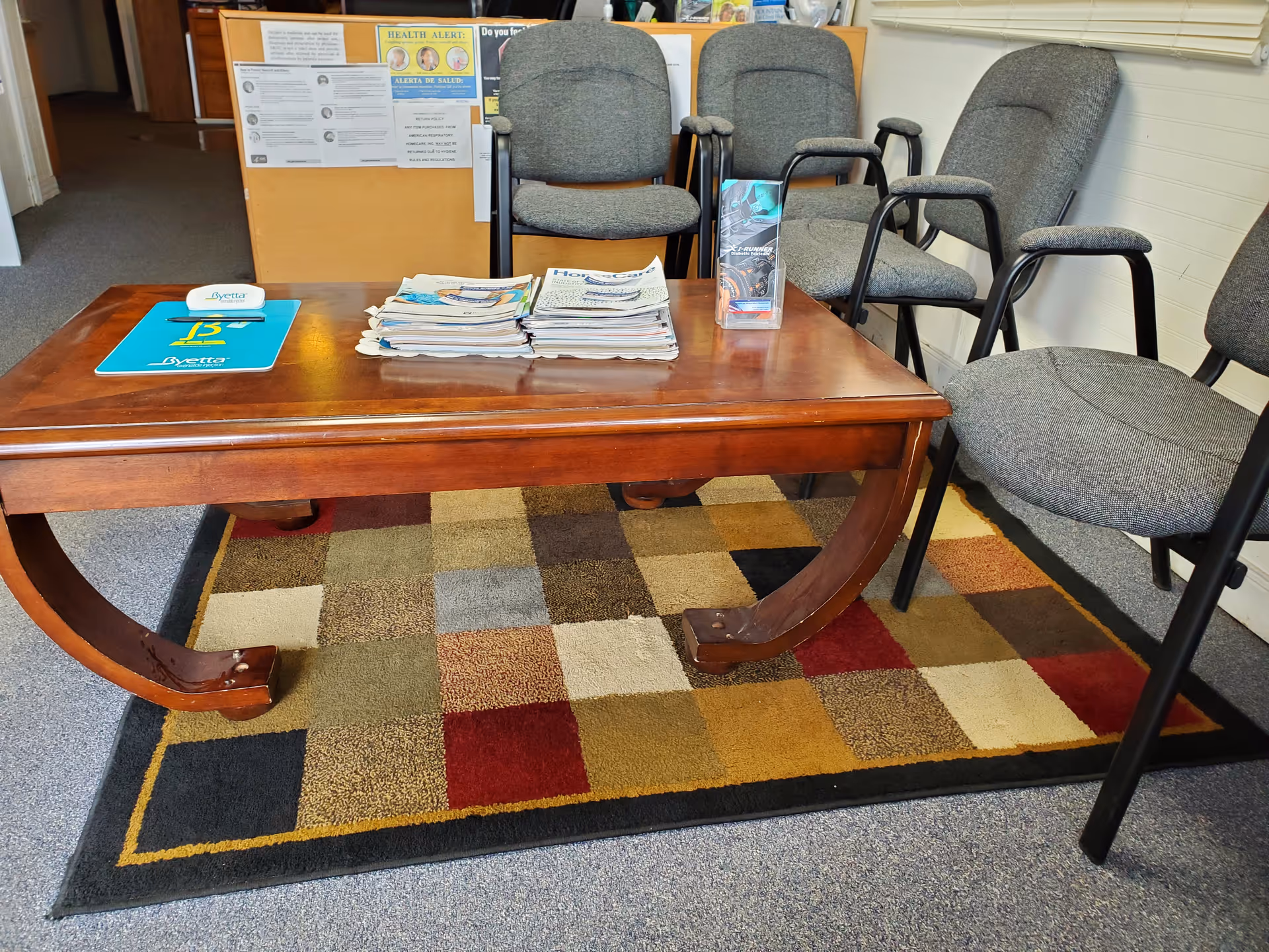 A wooden table holds stacked newspapers and brochures in a waiting area with chairs.