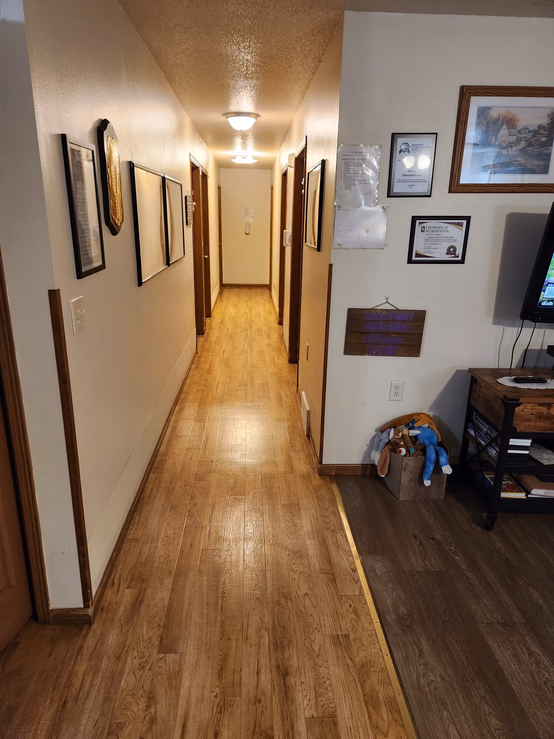 Narrow hallway in a senior living facility with wooden floors, framed art, and doors.