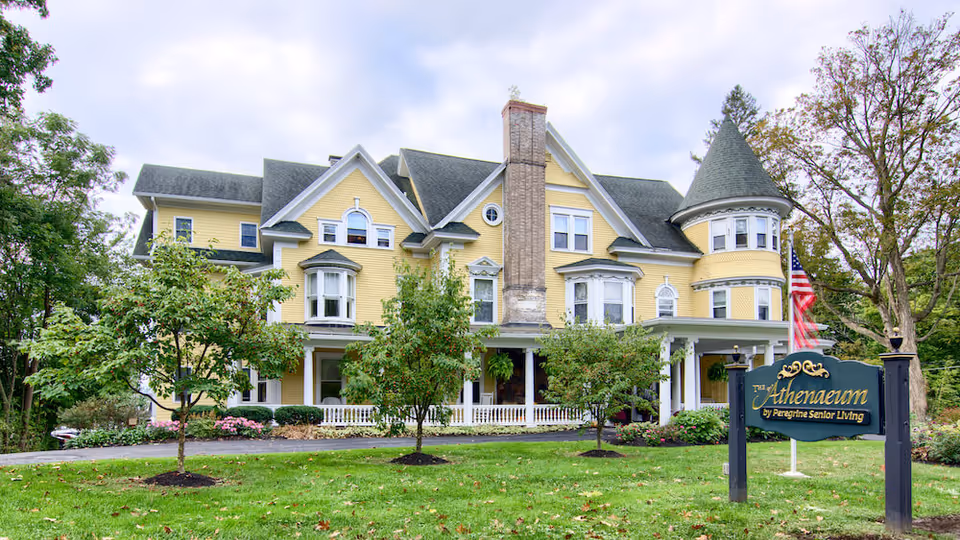 Yellow Victorian-style senior living facility front with a sign, trees, and a manicured lawn.