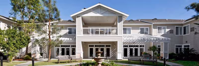 A two-story senior living center exterior with central entrance, balconies, trees, benches, and a fountain.