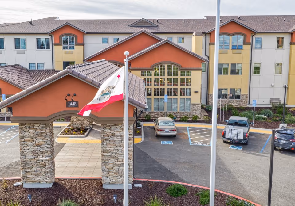 Front entrance of a multi-story senior living building with a stone-pillared porte-cochère, a flagpole flying the California flag, and parked cars in handicap spaces.