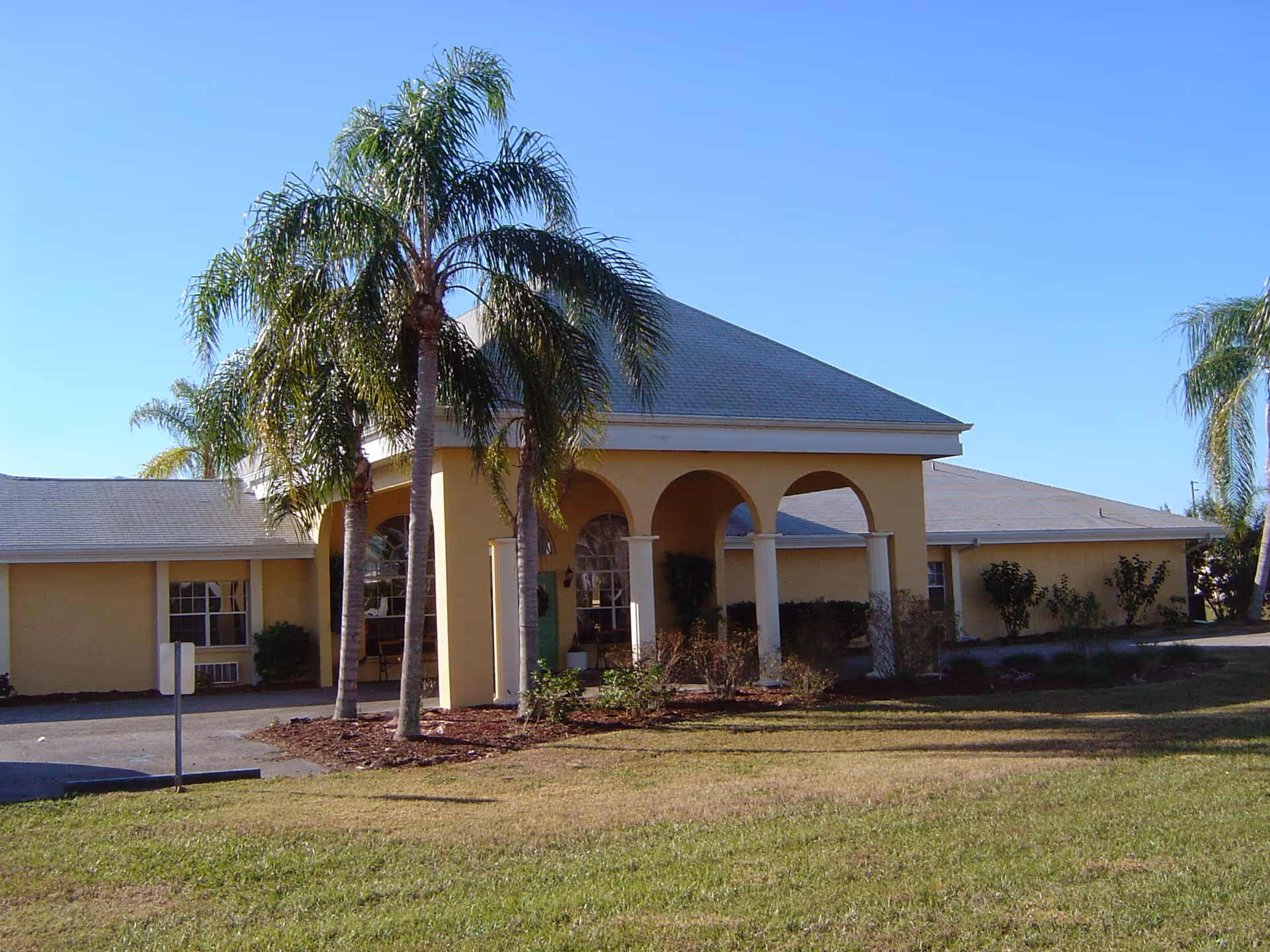 Sunny exterior view of Courtyard Retirement Center's front entrance with arches and palm trees.