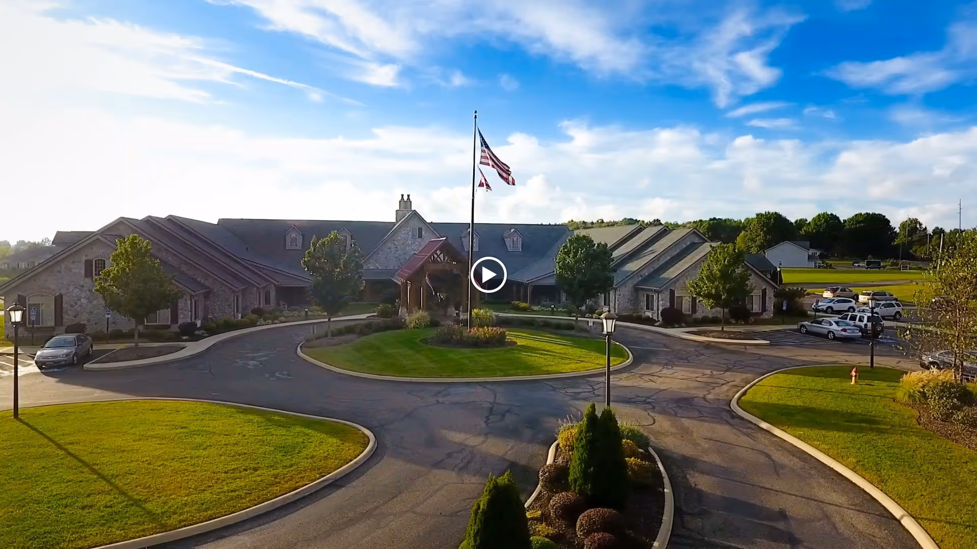 Front exterior of a senior living facility with a circular driveway, flagpole, and landscaped lawns under a blue sky.