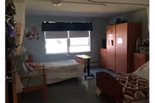 Shared senior living bedroom with two beds, dresser, window, and chairs.