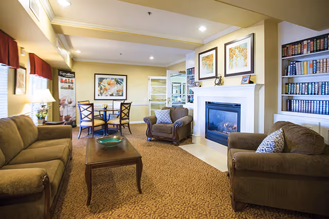 A cozy living room area in a senior living facility featuring a brown sofa, two armchairs with patterned cushions, a wooden coffee table with a decorative bowl, a fireplace with framed artwork above it, built-in bookshelves filled with books, and a small dining table with four chairs near the back wall.