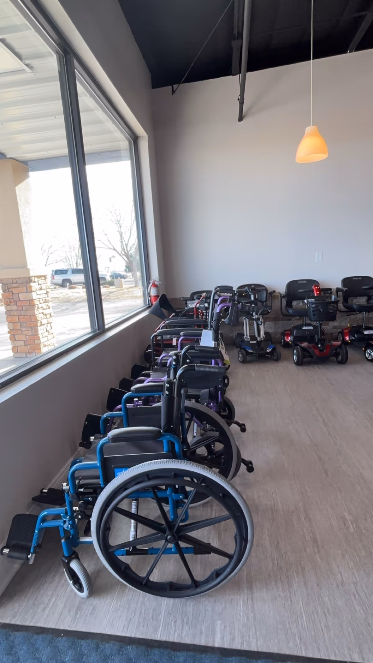Wheelchairs and mobility scooters lined up inside a bright facility hallway.